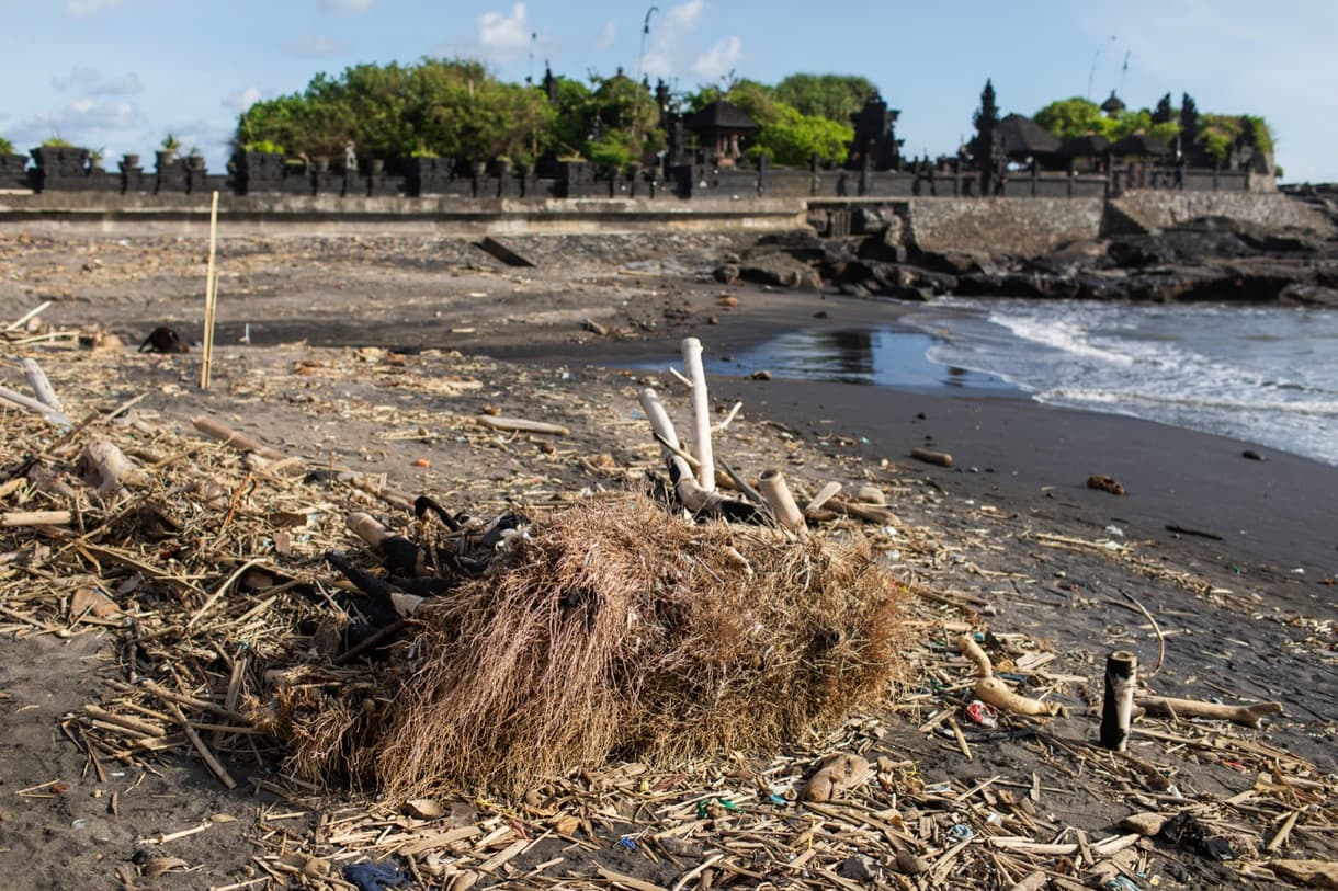 Plage de Tanah Lot, débris et pollution environnante sur le sable, avec temple en arrière-plan.