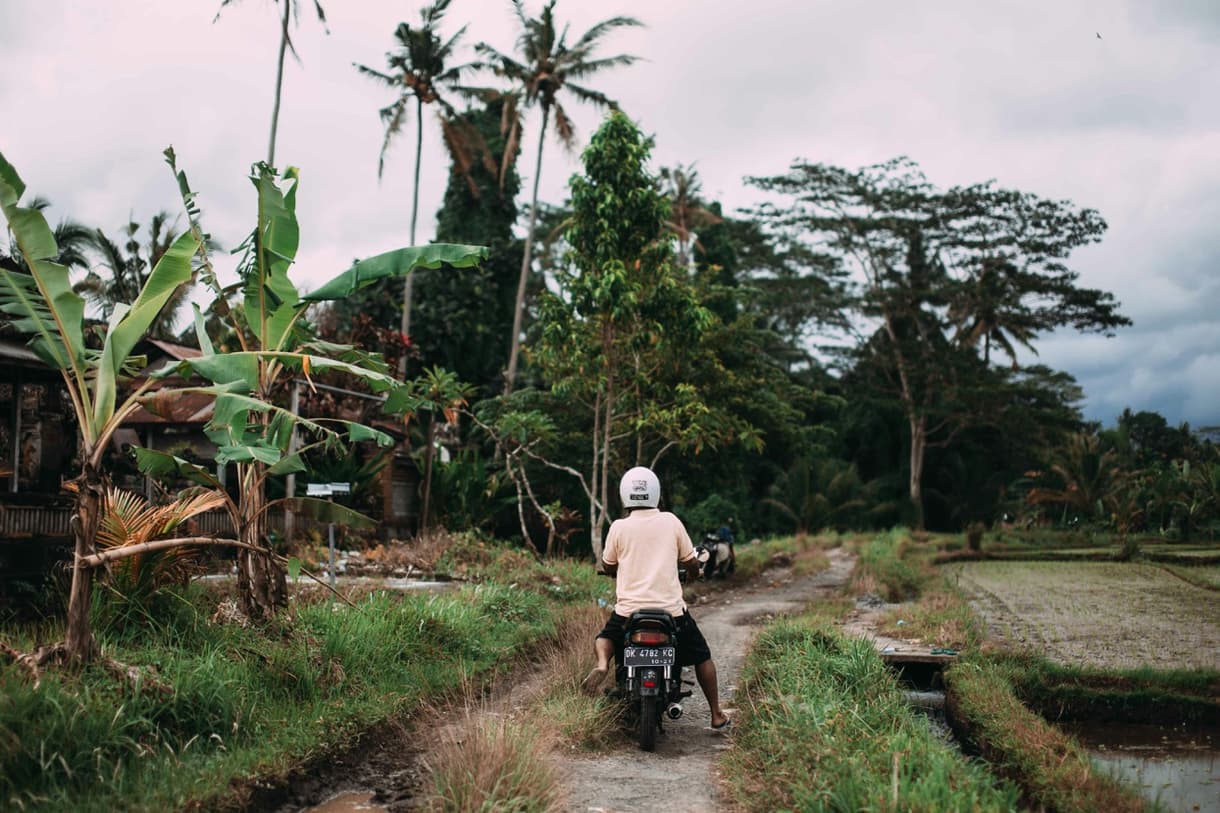 Un homme sur un scooter empruntant un chemin rural entouré de végétation tropicale à Bali