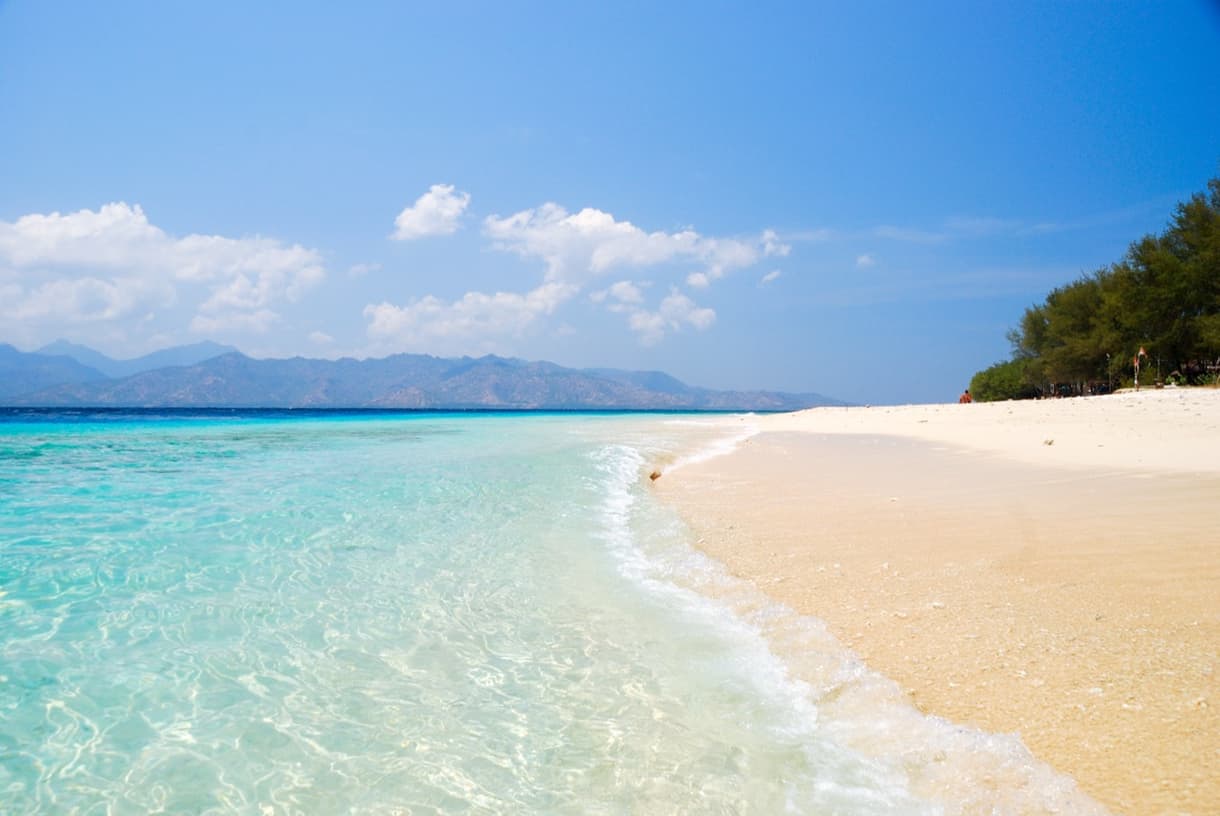 Plage de sable blanc bordée par une mer turquoise à Gili Air, avec des montagnes en arrière-plan.