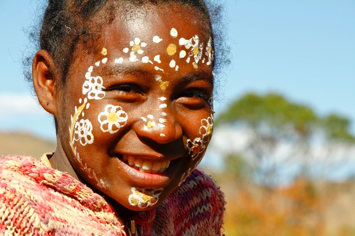 Enfant malgache avec le visage peint, affichant un sourire, sous un ciel bleu
