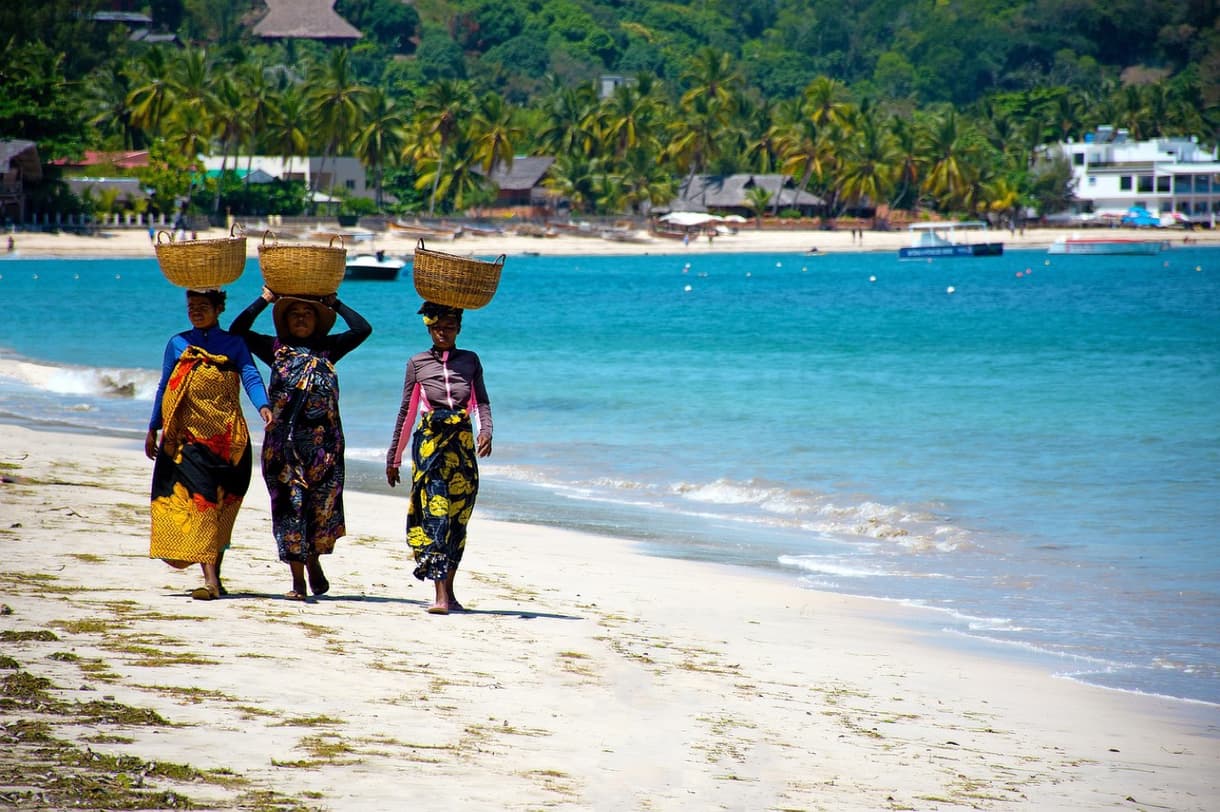 Trois femmes portant des paniers marchent sur une plage de Nosy Be, Madagascar, avec l'eau turquoise en arrière-plan.