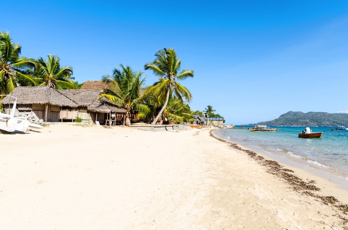 Plage de Nosy Komba avec maisons en paille et palmiers sous un ciel bleu