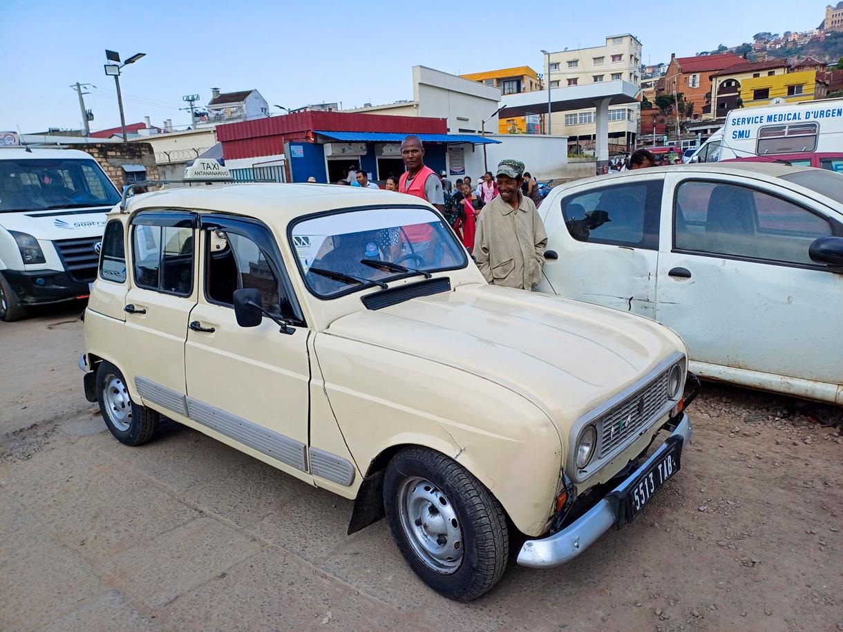 Taxi ancien sur une avenue de Madagascar, avec passants et bâtiments en arrière-plan.